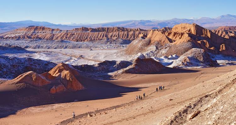 Visitantes caminando a través de un dramático paisaje lunar de color naranja y blanco con crestas erosionadas y llanuras en un inmenso valle desértico.