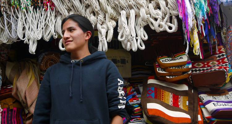 Vendedor entre bolsas tejidas de colores y textiles en un mercado andino.