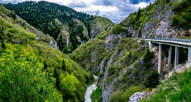 Canyon pittoresque avec une rivière sinueuse loin en dessous, des falaises boisées d'un vert dense et un haut pont avec un tunnel traversant le flanc de la montagne sous un ciel nuageux dramatique