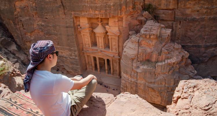 Una persona sentada al borde de un acantilado con vista a Petra.