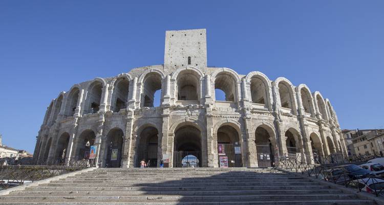 The well-preserved Roman amphitheatre of Arles standing grandly under a cloudless blue sky, viewed from its stone steps.