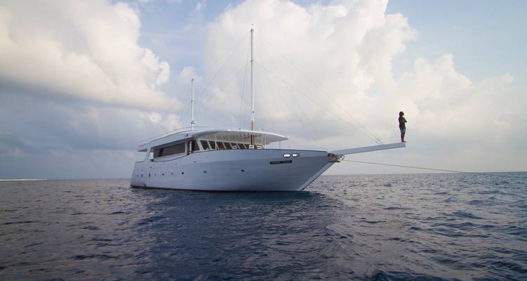 Une personne se tient sur le beaupré d'un yacht au-dessus d'une eau océanique calme et de nuages dramatiques.