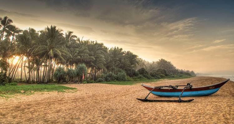 Empty beach with a traditional boat and palm trees at sunrise.