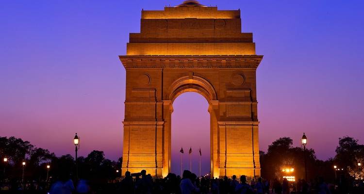 India Gate monument lit in golden lights against a vibrant purple dusk sky with crowds below.