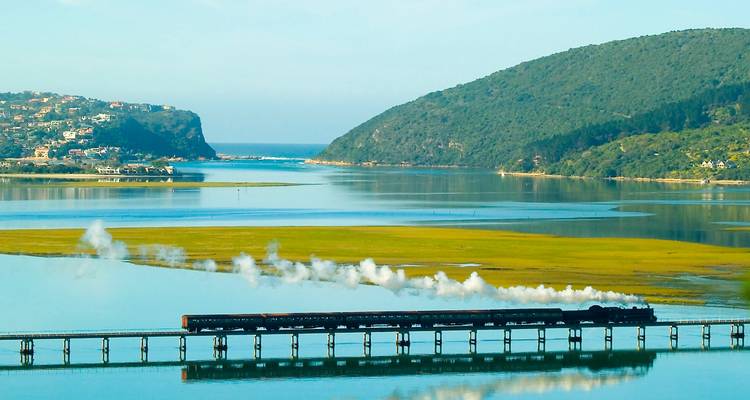 Train traversant un pont au-dessus d'un plan d'eau pittoresque avec des collines verdoyantes en arrière-plan.