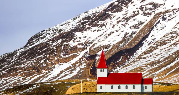 Église aux toits rouges se dressant contre une pente montagneuse abrupte aux traînées de neige sous un soleil éclatant.