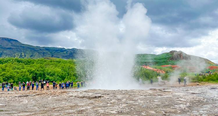 Puissante éruption d'un geyser projetant de la vapeur haut dans les airs tandis que les visiteurs se tiennent en large demi-cercle dans un parc géothermique.