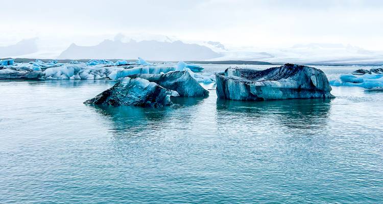 Lagon glaciaire bleu avec des icebergs épars flottant sous des ciels brumeux et des montagnes lointaines.
