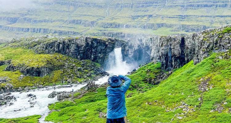 Photographe vêtu d'une veste bleue capturant une cascade brumeuse se précipitant dans une gorge rocheuse verdoyante.