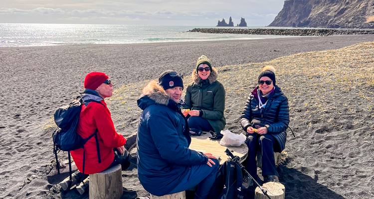 Quatre voyageurs vêtus de vêtements d'hiver sont assis sur des tabourets en bois et admirent la vue d'une plage de sable noir avec des formations rocheuses.