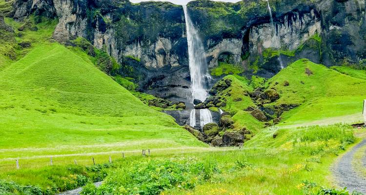 Une haute cascade en ruban se précipite d'une falaise couverte d'une mousse verte éclatante dans une vallée de prairie lumineuse.