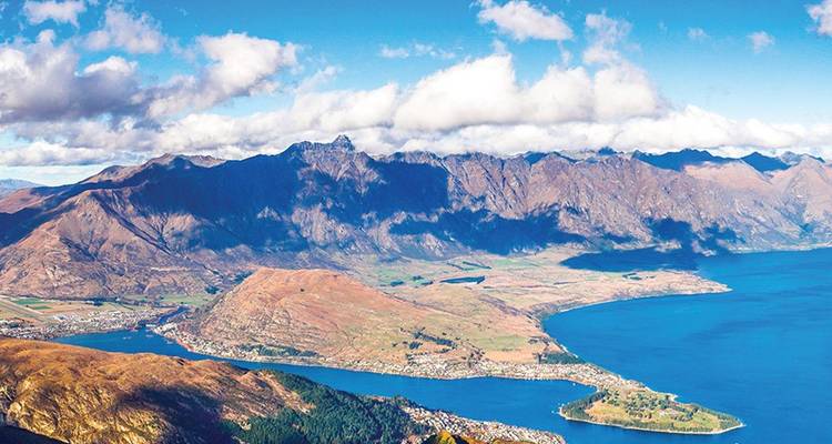 Sweeping aerial of lake and alpine mountains surrounding Queenstown with scattered clouds casting shadows.
