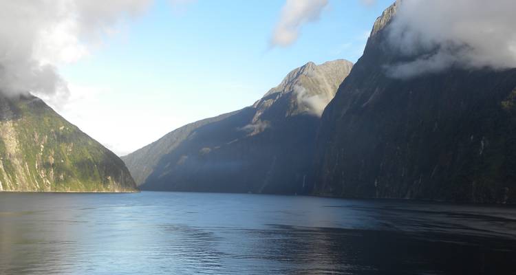 Steep forested cliffs enclose the calm, dark waters of Milford Sound under patches of cloud.