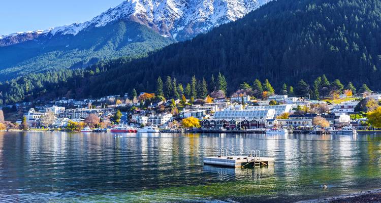 El pueblo junto al lago de Queenstown se refleja en las aguas tranquilas debajo de las laderas boscosas y una cumbre cubierta de nieve.