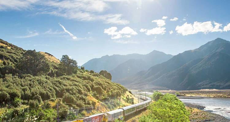 A long scenic passenger train winds through lush green hills toward towering mountains under a bright blue sky.
