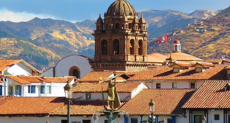 Rust-colored bell tower rising above red-tiled rooftops of Cusco with Andean hills in the background.