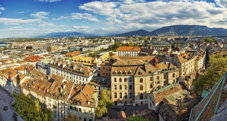 Aerial panorama of Geneva’s terracotta rooftops backed by distant alpine peaks and clouds.