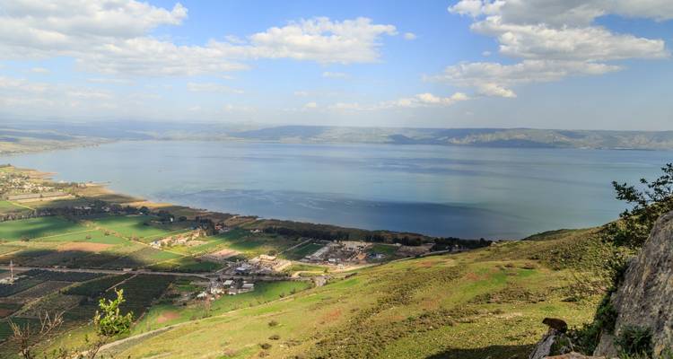 Serene expanse of the Sea of Galilee viewed from a green hillside under scattered clouds.