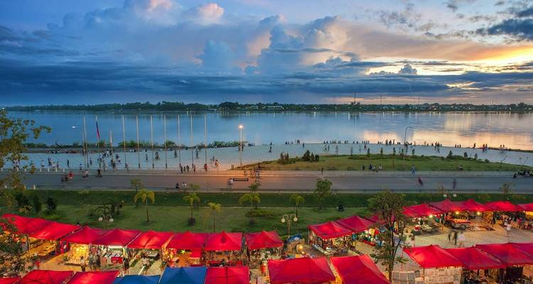 Night market by a river with colorful tents and distant skyline.