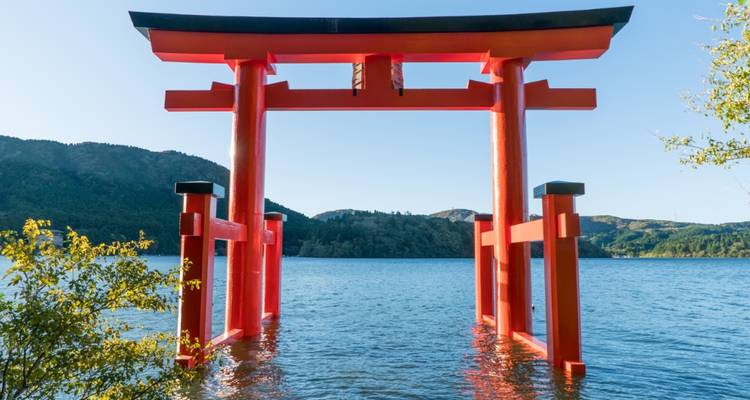 Portail torii rouge vif se dressant dans les eaux bleues du lac Ashi avec des collines boisées et un ciel dégagé en arrière-plan.