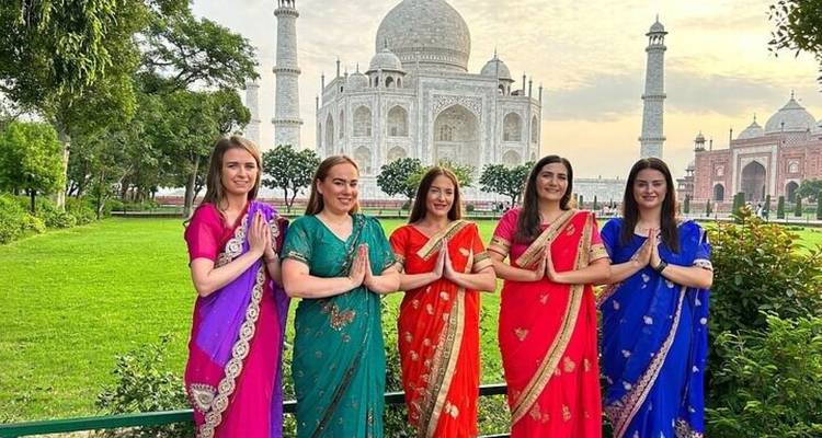 Five women in colorful saris posing with folded hands in front of the Taj Mahal at dawn.