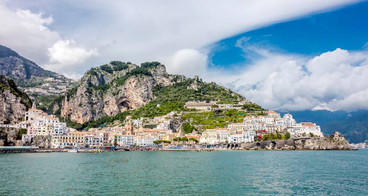 Vue panoramique en bord de mer de la ville d'Amalfi nichée sous des falaises verdoyantes escarpées et des nuages tourbillonnants.