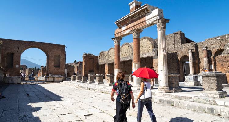 Deux touristes marchent main dans la main avec un parapluie rouge à travers les anciennes colonnes et les arcs de Pompéi sous un ciel bleu éclatant.