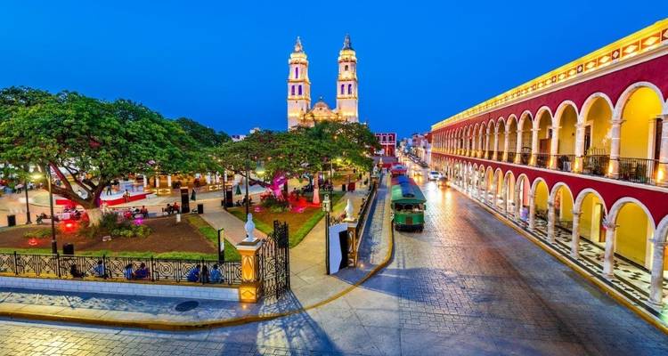 Plaza de la ciudad con una iglesia y árboles frondosos, iluminada en la tarde.