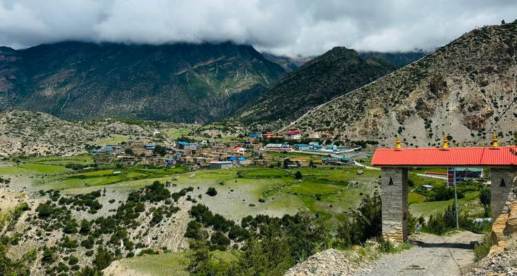 Stone gate overlooks a remote Himalayan village on a green plateau framed by rugged peaks and clouds.