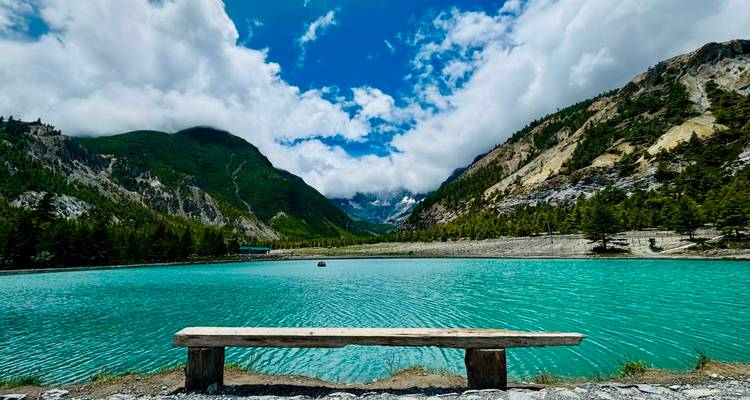 Turquoise alpine lake with a solitary wooden bench framed by dramatic forested mountains and clouds.