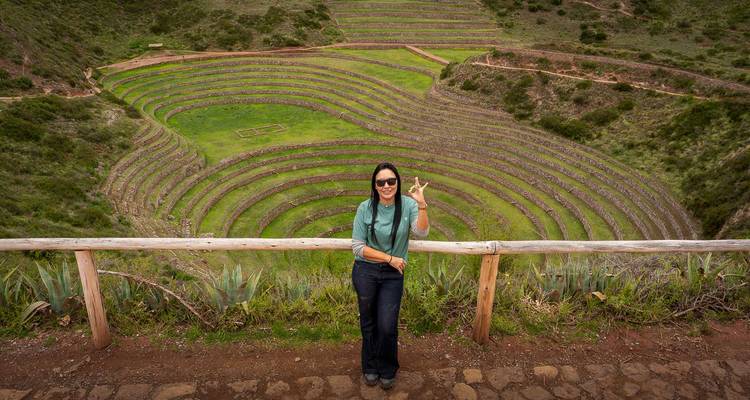Mujer sonriendo haciendo una señal de paz frente a las terrazas agrícolas circulares de Moray en el Valle Sagrado.