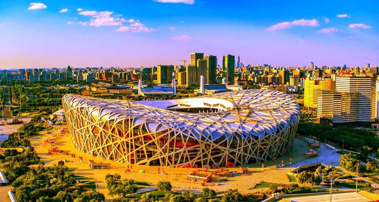 Panorama aéreo del Estadio Nido de Pájaro de Beijing rodeado por el horizonte de la ciudad bajo un cielo azul vibrante.