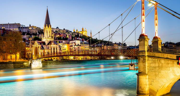 Un pont piétonnier illuminé enjambe la Saône à l'heure bleue, avec la vieille ville de Lyon qui brille en arrière-plan.