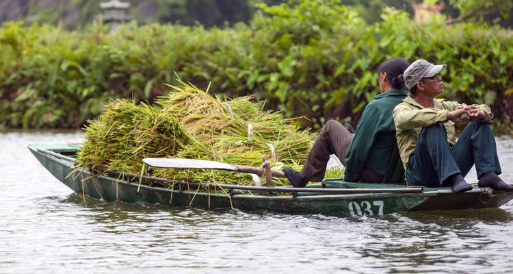 Dos personas sentadas en un bote lleno de plantas cosechadas en un río.