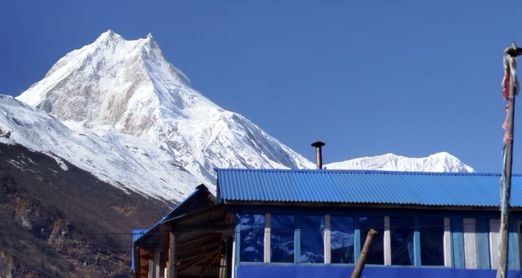 Pico nevado elevándose sobre una casa de té con techo azul en un pueblo himalayo