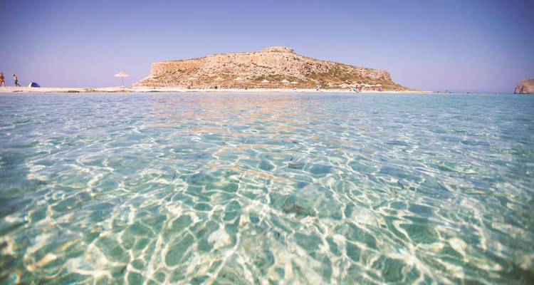 Eau claire avec une plage de sable et une colline rocheuse au loin.
