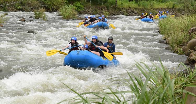 Meerdere blauwe boten storten zich door schuimende wildwaterstroomversnellingen omgeven door weelderige riviervegetatie.
