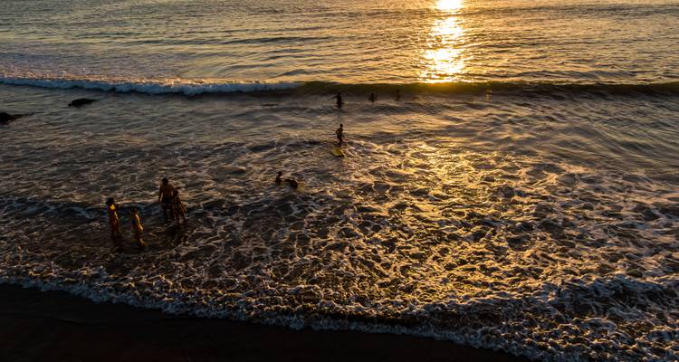Silhouetten van strandgangers spelen in zachte golven terwijl de zon ondergaat en gouden reflecties op het water werpt.