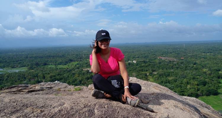 Woman sitting on rocky summit overlooking expansive green forest and horizon under partly cloudy skies.