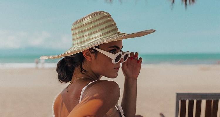 Stylish woman in wide-brimmed hat and sunglasses relaxing on a sandy tropical beach with turquoise sea backdrop.