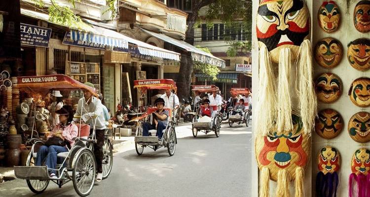 Translation from English to French:
**Promenades en cyclo-pousse et masques culturels exposés dans une rue animée.**
Note: "Cyclo" refers to cycle rickshaws (cyclo-pousse in French), which are common forms of transportation in some Asian countries, particularly Vietnam.