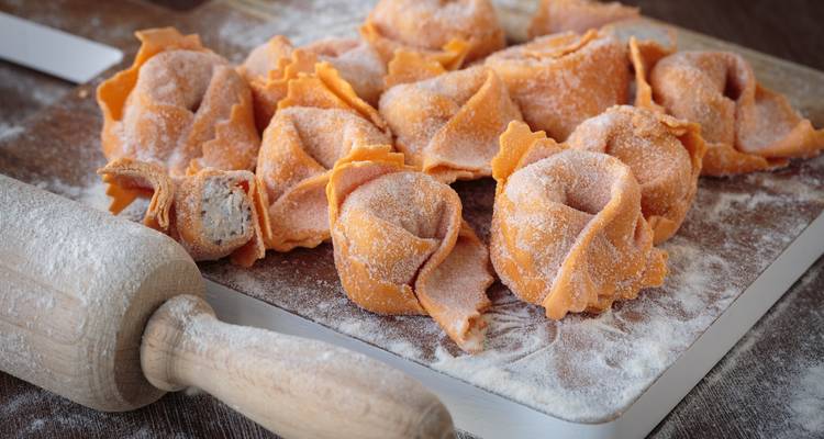 Tortellini de naranja recién hechos espolvoreados con harina en una tabla de madera junto a un rodillo.