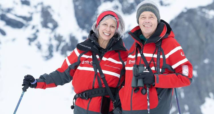 Glimlachend volwassen stel in rode expeditiejassen staat op sneeuw met trekkingstokken tegen een rotsachtige Antarctische achtergrond.