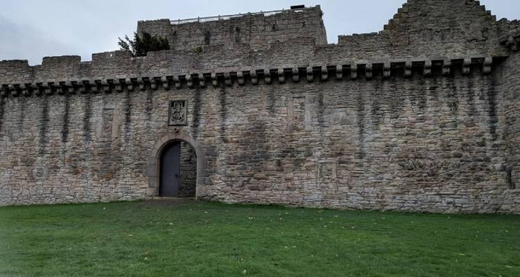 Steinerne Burgmauer mit einem Eingangstor auf einer Wiese.