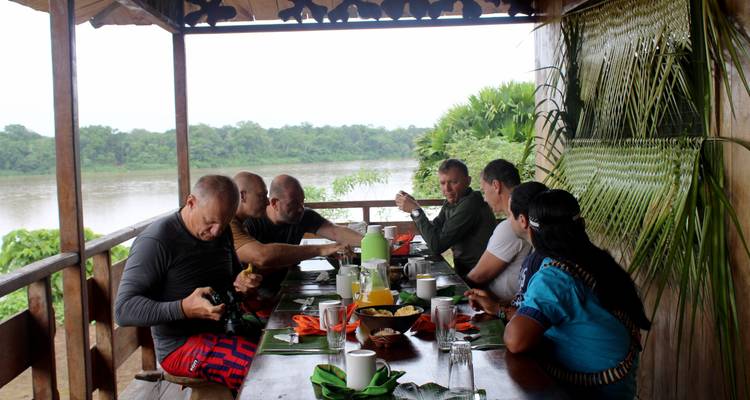 Personas cenando en una mesa en una terraza frente al río.