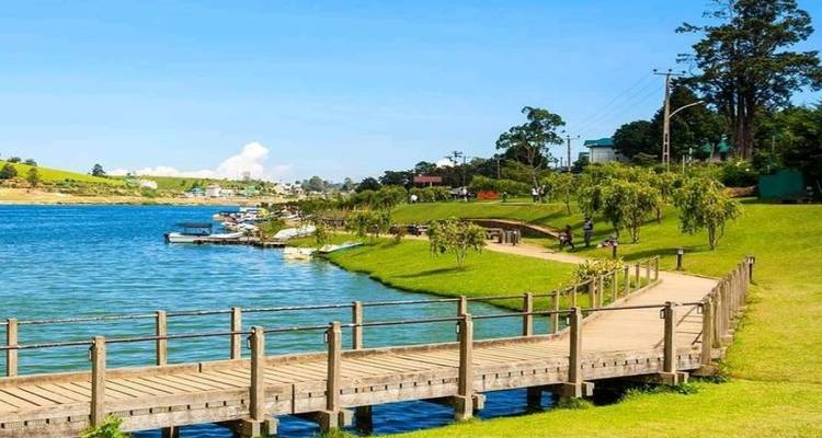Sunny lakeside scene with a wooden boardwalk curving along turquoise water, green lawns and distant boats.