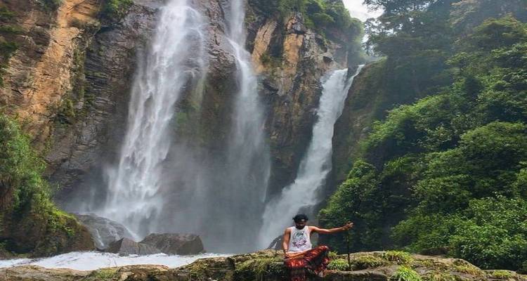 Traveler standing on rocks in front of a tall, misty waterfall cascading down forested cliffs.