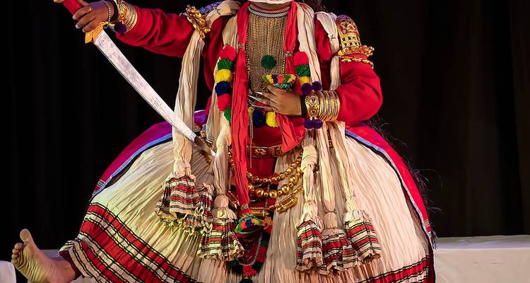 Artiste de Kathakali avec maquillage dramatique et épée sur scène.