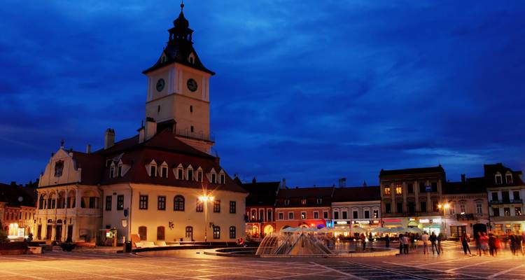 La Place du Conseil de Brasov à l'heure bleue avec la tour de l'ancien hôtel de ville éclairée contre un ciel bleu profond.