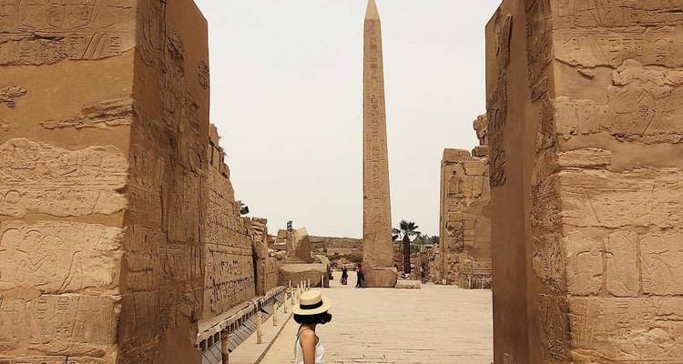 Een vrouw die naar een hoge obelisk loopt, omringd door oude ruïnes.